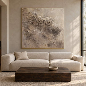 In a living room above seating, an artwork with a gold-colored frame shows irregular beige and taupe relief-like textures on a pale square ground.