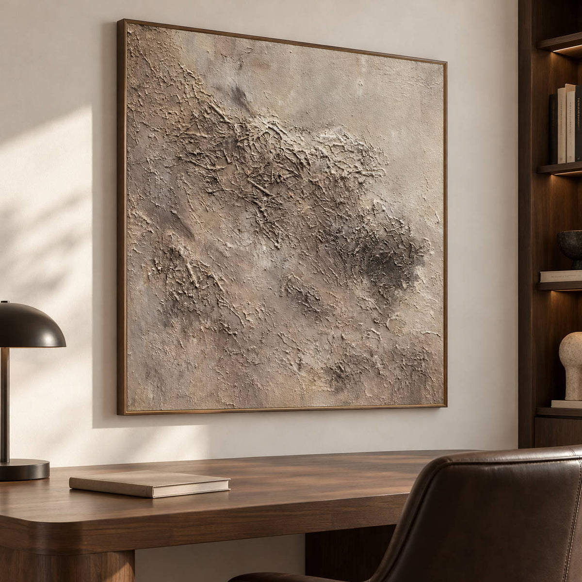 Irregular beige and taupe relief-like textures on a pale square ground in an artwork with a brown wood-look frame, displayed in a study with a desk.
