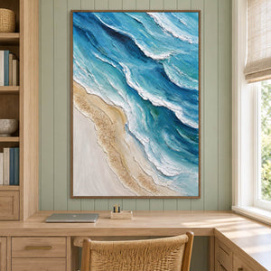 Layered blue-green waves with raised white edges beside a pale sandy shore in an artwork with a brown wood-look frame, displayed in a study with a desk.